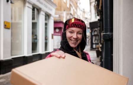 Téléchargez les photos : Smiling bike courier woman delivering a parcel at the door in a narrow street. She is wearing cap, sunglasses and hood. - en image libre de droit