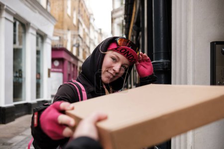 Téléchargez les photos : Portrait d'une passagère à vélo livrant un colis à porte plate dans une rue étroite de Londres. La main du client prend le colis. - en image libre de droit