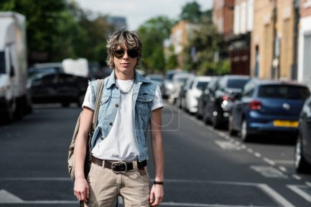 Foto de Una persona andrógina mirando una cámara en un camino de Londres. Hay un montón de coches aparcados y es un día soleado. - Imagen libre de derechos