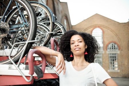 Photo for Portrait of black woman leaning on bike rack. She is looking at camera smiling. Green transportation and security concept. - Royalty Free Image