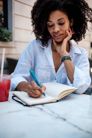 Lächelnde Frau, die auf einem Terrassentisch im Freien ihr Tagebuch schreibt. Es ist Sommer und es steht ein Saftbecher auf dem Tisch. Technikfreie Hobbys und Literaturkonzept.