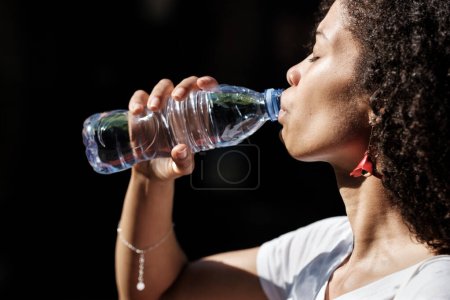 Foto de Atractiva mujer negra bebiendo una botella de agua en un caluroso día de verano. Concepto hidratante natural. - Imagen libre de derechos
