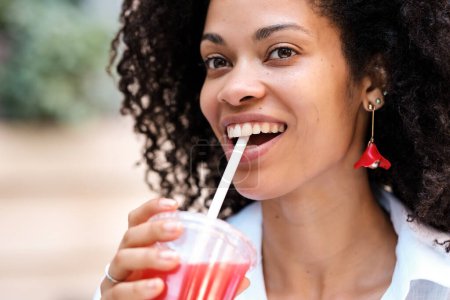 Foto de Mujer negra feliz bebiendo un jugo de fresa con una pajita. Ella sonríe y mira hacia otro lado. Retrato de cerca. concepto de estilo de vida saludable. - Imagen libre de derechos