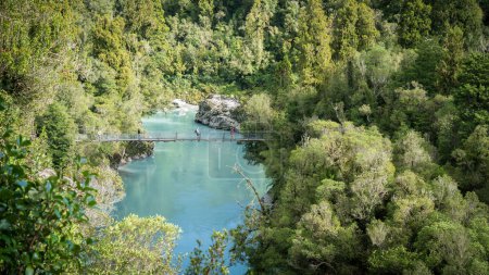 Menschen überqueren Schwingbrücke über Schlucht mit azurblauem Wasser, umgeben von dichtem Wald, Neuseeland.