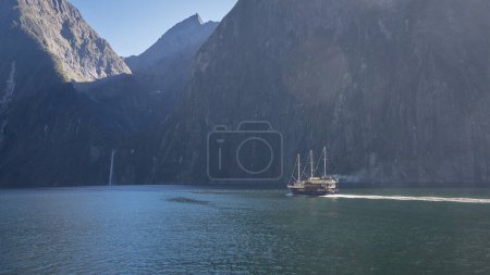 Photo for Boat sailing through fjord with steep rocky walls and waterfall in distance, Fiordland, New Zealand. - Royalty Free Image