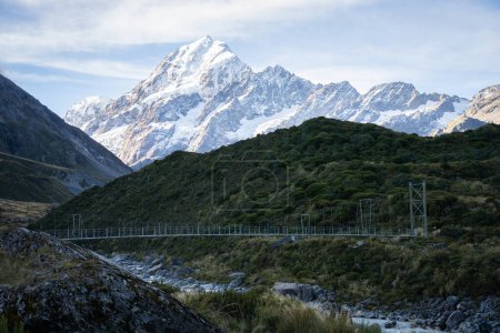 Swing bridge leading through alpine valley with huge snowy mountain behind it, Mt Cook, New Zealand.