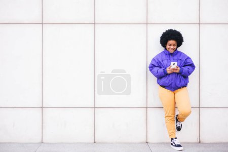 Foto de Joven mujer latina afro de pie en la pared blanca usando el teléfono y sonriendo, espacio para la copia. Foto de alta calidad - Imagen libre de derechos