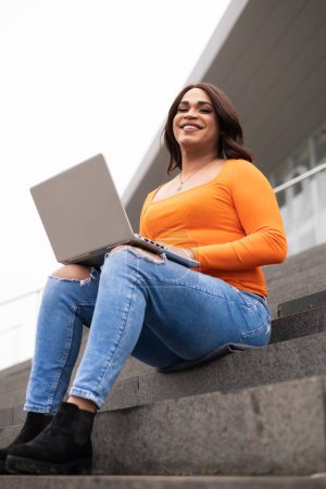 Photo for Close-up portrait of trans latin woman using laptop in campus stairs. She is smiling. Trans inclusion concept. High quality photo - Royalty Free Image