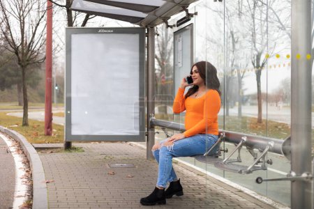 Full view of trans latin woman sitting in bus stop and using mobile phone. Copy space. High quality photo