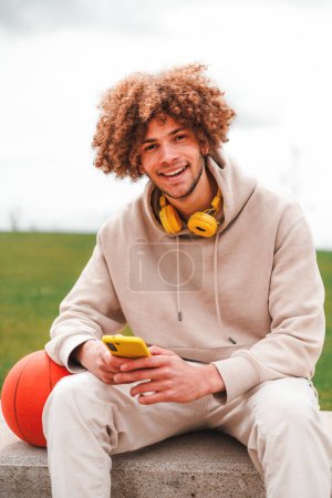 Hombre pelo rizado sosteniendo teléfono inteligente y sonriendo a la cámara sobre un verde leavpark es fondo. Imágenes FullHD de alta calidad