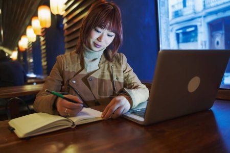 Foto de Mujer asiática trabajando con su portátil mientras escribe en su cuaderno. - Imagen libre de derechos