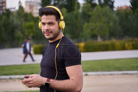 Photo for Smiling latin millennial sportsman in headphones choosing music on mobile phone for running and training outdoors, cheerful hipster guy holding smartphone for select music - Royalty Free Image