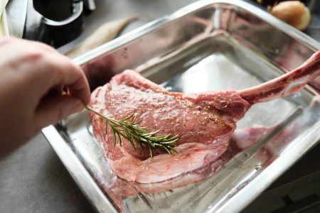 Téléchargez les photos : Gros plan de la main de l'homme mettant du romarin sur la viande d'agneau avec des épices - en image libre de droit