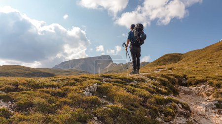 Foto de A man with a big hiking backpack hiking on a plateau in high Italian Dolomites. There are golden mountain slopes around the pathway. High and sharp mountains around. Remote and raw area. Calmness - Imagen libre de derechos