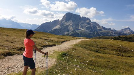 Foto de A woman with hiking backpack hiking along a gravelled road in Italian Dolomites. There is a massive mountain chain in front of her, she is enjoying the trek. High Alpine plateau with lush green meadow - Imagen libre de derechos