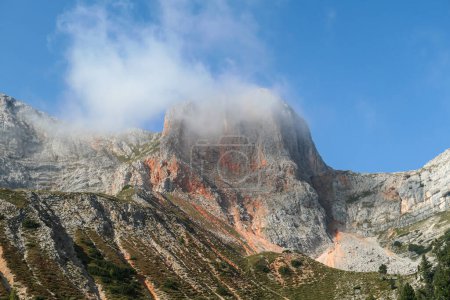 Foto de Una vista de cerca de un pico de montaña distante en Dolomitas italianas. La montaña tiene un color rojo distintivo, mezclándose con los lados grises. En las estribaciones hay un denso bosque. Hay una nube delgada en frente - Imagen libre de derechos