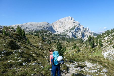 Foto de Una mujer con una mochila de senderismo senderismo en un camino angosto en alta Dolomitas italianas. Cadena de montaña empinada y afilada en la parte posterior. Está cruzando un exuberante prado verde. Descubrir y explorar la naturaleza - Imagen libre de derechos