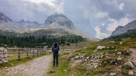 Téléchargez les photos : Un homme avec un grand sac à dos de randonnée le long d'une route gravelée dans les Dolomites italiennes. Il y a une clôture en bois le long de la route. Chaînes de montagnes acérées et pierreuses devant. Quelques rochers sur la prairie verte. - en image libre de droit
