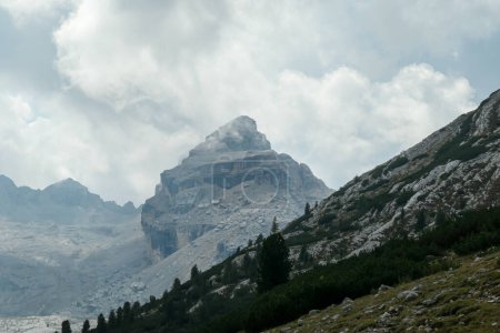 Téléchargez les photos : Vue rapprochée sur une montagne pierreuse et escarpée dans les Dolomites italiennes. La montagne massive est entourée de nuages. Il y a une pente verte d'une autre montagne en face. Zone éloignée et désolée. Couvert - en image libre de droit
