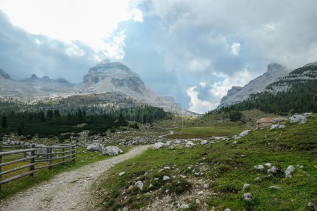 Téléchargez les photos : Une vue panoramique sur une vallée avec une route gravelée le long de celle-ci dans les Dolomites italiennes. Il y a une clôture en bois le long de la route. Chaînes de montagnes acérées et pierreuses devant. Quelques rochers sur la prairie verte. - en image libre de droit