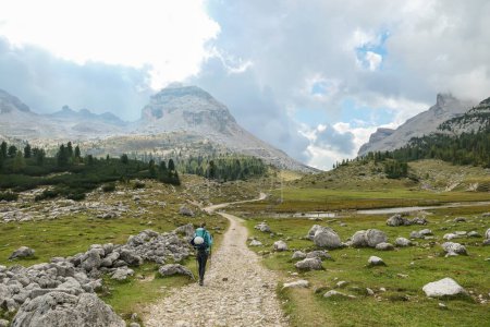 Téléchargez les photos : Une femme avec un sac à dos de randonnée randonnée sur une route gravelée dans les hautes Dolomites italiennes. Il y a une forêt dense au pied de la colline, et une chaîne de montagnes raide et forte à l'arrière. Découvrir et explorer - en image libre de droit