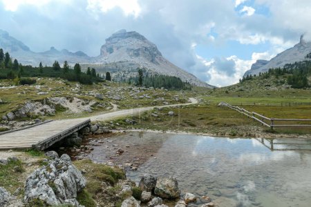 Téléchargez les photos : Un petit étang sous un pont en bois dans les Dolomites italiennes. L'étang reflète les montagnes environnantes et le ciel. Chaînes de haute montagne à l'arrière. Forest sur le côté. Région déserte et éloignée - en image libre de droit
