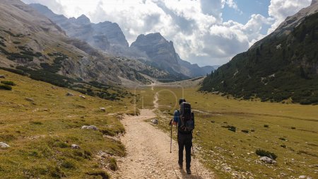 Téléchargez les photos : Un homme avec un sac à dos de randonnée randonnée sur une route gravelée dans les hautes Dolomites italiennes. Il y a une forêt dense au pied de la colline, et une chaîne de montagnes raide et forte à l'arrière. Découvrir et explorer - en image libre de droit
