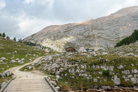 Téléchargez les photos : Une route gravelée menant à travers un petit pont en bois à un chalet dans les Dolomites italiennes. Hautes montagnes pointues derrière le chalet. Prairie avec de nombreux rochers devant. Endroit isolé et désolé. - en image libre de droit