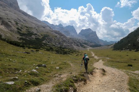 Téléchargez les photos : Une femme avec un sac à dos de randonnée randonnée sur une route gravelée dans les hautes Dolomites italiennes. Il y a quelques arbres sur les parties inférieures, et une chaîne de montagnes raide et forte à l'arrière. Découvrir et explorer - en image libre de droit