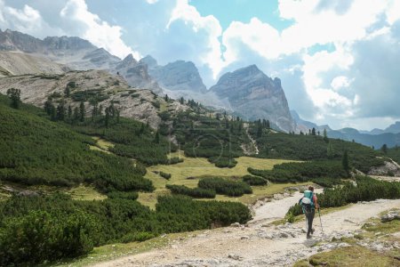 Téléchargez les photos : Une femme avec un sac à dos de randonnée randonnée sur une route gravelée dans les hautes Dolomites italiennes. Il y a quelques arbres sur les parties inférieures, et une chaîne de montagnes raide et forte à l'arrière. Découvrir et explorer - en image libre de droit
