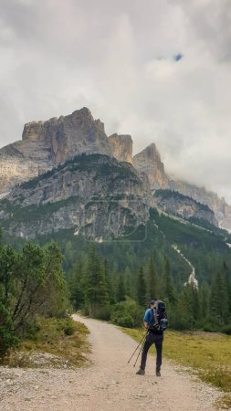 Téléchargez les photos : Un homme avec un sac à dos de randonnée pédestre sur un sentier en gravier dans les hautes Dolomites italiennes. Il y a quelques arbres sur les parties inférieures, et une chaîne de montagnes raide et pointue à l'arrière. Découvrir et explorer - en image libre de droit