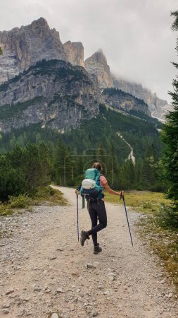 Téléchargez les photos : Une femme avec un sac à dos de randonnée pédestre sur un sentier en gravier dans les hautes Dolomites italiennes. Il y a quelques arbres sur les parties inférieures, et une chaîne de montagnes raide et pointue à l'arrière. Découvrir et explorer - en image libre de droit