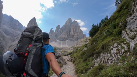 Téléchargez les photos : Un homme avec un grand sac à dos prenant un selfie tout en marchant le long d'un étroit sentier dans les Dolomites italiennes. Il y a une montagne massive avec des pentes très raides et pointues. Des montagnes plus petites autour. Paysage brut - en image libre de droit