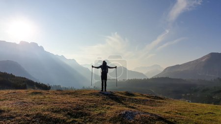 Photo for A man with hiking sticks spreading his arms wide open on an early morning in Italian Dolomites. The valley below is shrouded in morning haze. In the back there are high mountain chains. Golden hour - Royalty Free Image