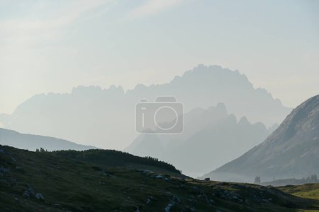 Foto de An early morning in Italian Dolomites. Lush green meadow in front. The valley is shrouded in morning haze. In the back there are high mountain chains. Sun slowly rising above the peaks. Golden hour - Imagen libre de derechos