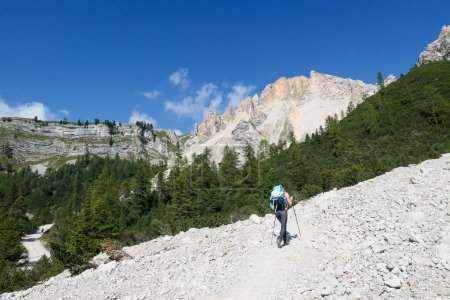 Téléchargez les photos : Une femme avec un sac à dos de randonnée randonnée sur une route gravelée dans les hautes Dolomites italiennes. Il y a une forêt de dence au pied de la colline, et une chaîne de montagnes escarpée et forte à l'arrière. Découvrir et explorer - en image libre de droit