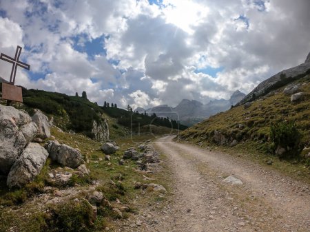 Téléchargez les photos : Une route gravelée qui traverse les Dolomites italiennes. Il y a une croix métallique sur le rocher à côté de la route. De hautes montagnes autour. Les pentes sont vertes. Couvert. Spiritualité et calme. - en image libre de droit