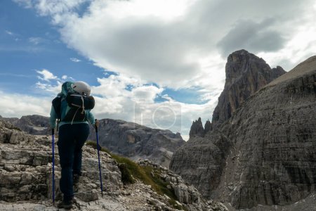 Photo for Woman with big backpack and sticks hiking in high Italian Dolomites. She goes up a steep slope. There are many sharp peaks in front of her. She is going up. There are a few trees around. Sunny day. - Royalty Free Image