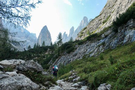 Foto de Una mujer con una gran mochila de senderismo senderismo en alta Dolomitas italianas. Hay muchos picos afilados detrás. Ella camina por un sendero muy estrecho. Hay algunos árboles alrededor. Día soleado. Ejercicio al aire libre - Imagen libre de derechos