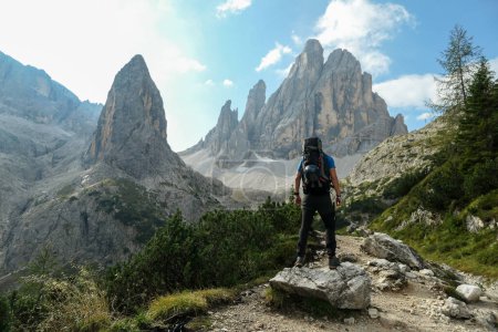 Foto de Un hombre con una gran mochila de senderismo en alta Dolomitas italianas. Hay muchos picos afilados detrás. Él está de pie sobre una gran roca, disfrutando de la vista. Hay algunos árboles alrededor. Día soleado. Exterior - Imagen libre de derechos