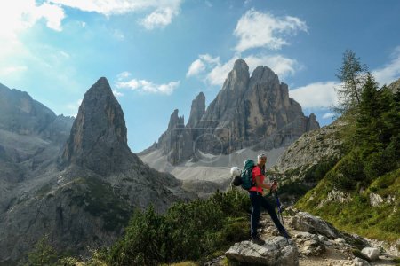 Photo for A woman with big hiking backpack hiking in high Italian Dolomites. There are many sharp peaks behind her. She is enjoying the hike, smiling. There are a few trees around. Sunny day. Outdoor exercising - Royalty Free Image