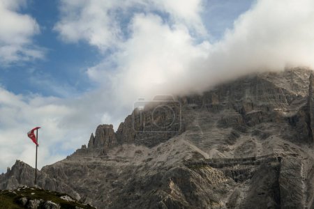 Foto de Una bandera tirolesa ondeando un viento en los Dolomitas italianos. Hay muchos picos altos y agudos cubiertos de nubes claras. Deslizamientos de tierra peligrosos, con muchas piedras perdidas alrededor. Paisaje crudo y virgen. - Imagen libre de derechos