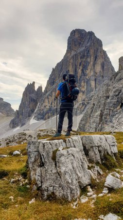 Photo for Man with big backpack and sticks, hiking in high Italian Dolomites. There are many sharp peaks in front of him. He is standing on a big boulder. Lots of lose stones and landslides. Sunny day. Outdoor - Royalty Free Image