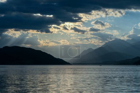 Photo for A sunset by Millstaetter lake in Austria. The lake is surrounded by high Alps. Calm surface of the lake reflecting the sunbeams. The sun sets behind the mountains. Thick clouds above. Natural beauty - Royalty Free Image