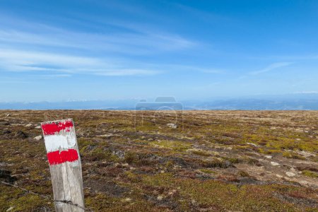 Foto de A red-white-red path mark on a wooden fence. A vast, golden pasture on top of Sauofen, Austrian Alps. A lot of big stones on the grounds. High mountain chains in the back. Blue sky, with a few clouds. - Imagen libre de derechos