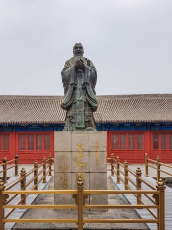 Téléchargez les photos : A statue of Confucius in front of Confucius temple in Beijing, China. The stature of the philosopher is surrounded by golden railing. There is a red pavilion in the back. Tradition and cult place - en image libre de droit