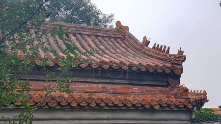 Foto de A close up on the rooftop of a pavilion in Forbidden City in Beijing, China. The roof has orange tiles and decorative small figures of guardians at the edges. Overcast. A few tree branches on the side - Imagen libre de derechos