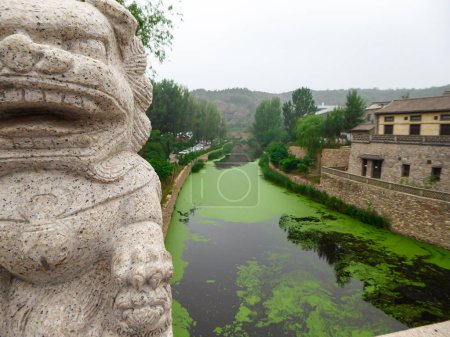 Photo for A view on the river in a small village in nothern China. The river is overgrown with green plankton. there is a lion figure on the bridge. Houses on both sides of the river. Serenity and calmness - Royalty Free Image