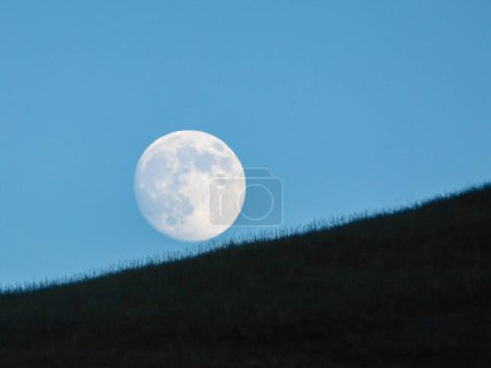 Photo for A full moon rising above the hilly grassland in the nearby of Xilinhot, Inner Mongolia, China. The sky behind it is cloudless and light blue. Rounded face of the moon. New moon. Calmness - Royalty Free Image