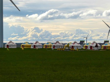Foto de Yellow and white traditional yurts located on a pasture in Xilinhot in Inner Mongolia. Endless grassland. Blue sky with a few thick clouds. Wind turbines in the back. Clean energy. Nomadic way of life - Imagen libre de derechos